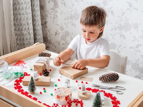 Kid wraps handmade Christmas presents in craft paper with colorful pompons and snowflake ribbons. Child prepares gifts for New Year celebration. Peaceful leisure activity before winter holidays.