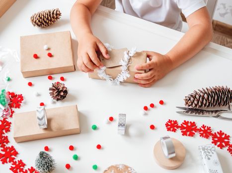 Kid wraps handmade Christmas presents in craft paper with colorful pompons and snowflake ribbons. Child prepares gifts for New Year celebration. Peaceful leisure activity before winter holidays.