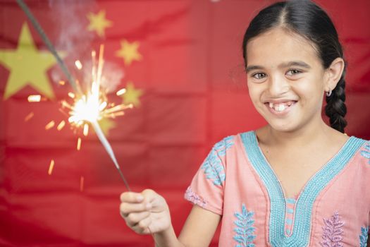 Smiling Young Girl Kid holding Sparkler in hand with Chinese flag as background - concept showing Celebration of Chinas National or Republic Day.