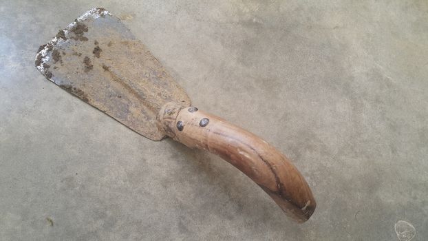 Closeup of old style garden trowel with metallic blade and wooden handle placed over a concrete floor. Trowel is old, rugged and dirty with mud.