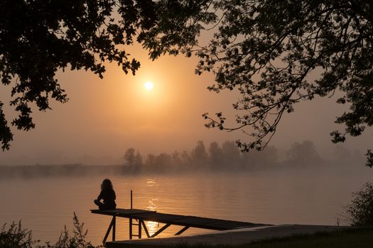 silhouette girl at wooden jetty during sunset in the early morning over the river maas in limburg in holland with the trees mist and hazy fog