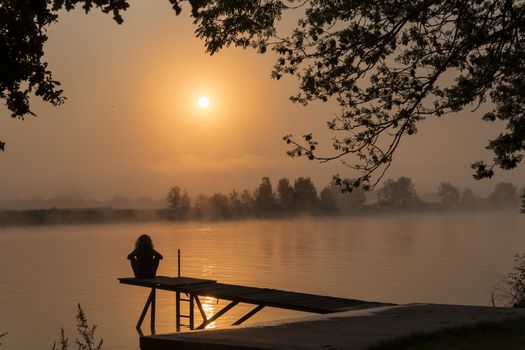 silhouette girl at wooden jetty during sunset in the early morning over the river maas in limburg in holland with the trees mist and hazy fog