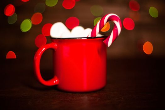 Cocoa in red mug with marshmallows and candy cane on dark wooden background with bokeh lights