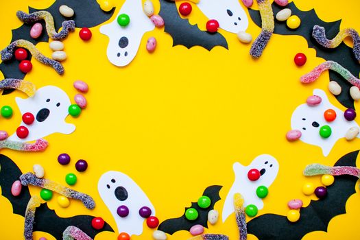 frame made of paper homemade bats and paper ghosts and of multicolored candies and worms from gummy on a bright yellow background