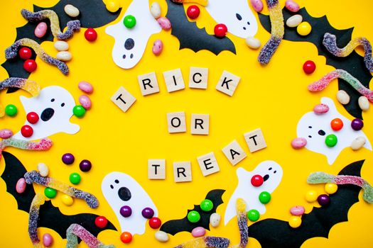 inscription from wooden blocks trick or treat and frame made of paper homemade bats and paper ghosts and multicolored candies and worms from gummy on a bright yellow background