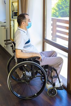 Lonely young man wearing face mask sitting in a wheelchair alone looking out the window. Focus on his face.