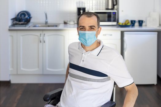 Smiling Young Handicapped Man Sitting On Wheelchair In Kitchen.  Young man wearing face mask sitting infront of kitchen. Focus on his face.