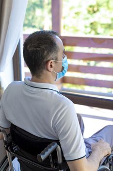 Lonely young man wearing face mask sitting in a wheelchair alone looking out the window. Focus on his head.