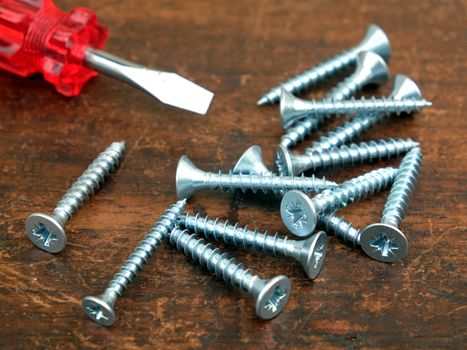 Some many silver screws and small handy screw driver on brown wooden surface side view closeup view