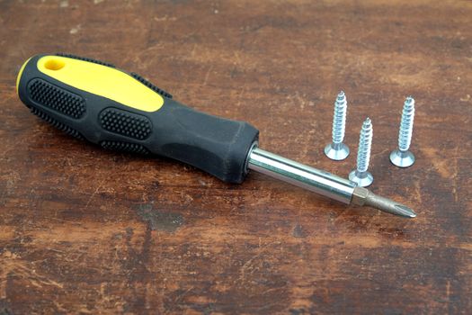 Handy screw driver and three sharp screws on brown wooden background closeup view