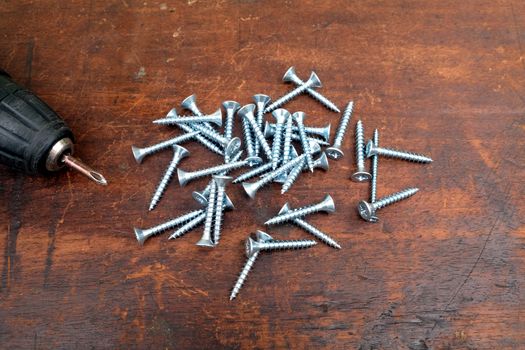 Heap of many light gray screws and old motor screw driver on brown wooden background top view closeup view