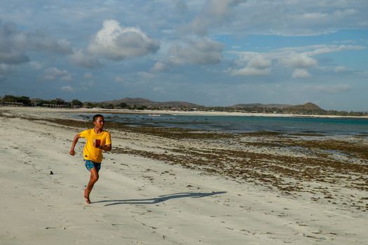 Running man jogging on beach. Indonesian teenager in shorts and a yellow T-shirt running along the beach. Young athlete trains by the sea. Fit young male sport fitness model exercising in summer.