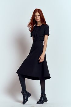 A woman in a black dress on a light background and pantyhose shoes red hair and pose in full growth. High quality photo