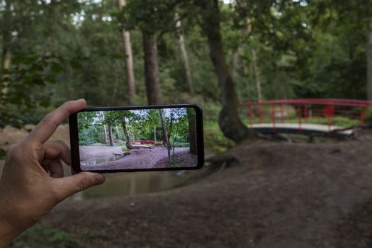 woman making a photo with smartphone of the  typical red japanese bridge in garden with forest trees and plants and sand at the border of small water