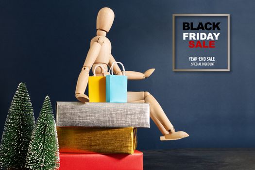 Black Friday sale, wooden doll sitting on gift box with shopping bag and christmas tree