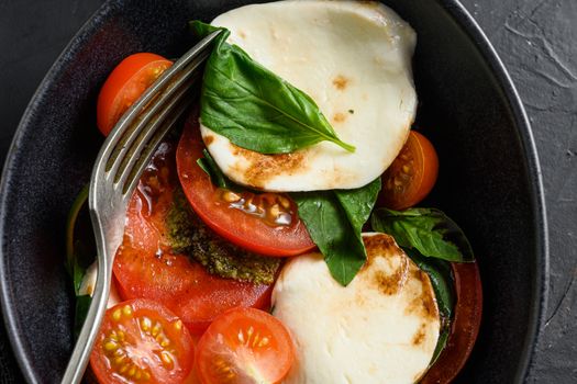 Cherry tomatoes, mozzarella buffalo cheese, basil, pesto sauce, and spices on black slate stone chalkboard. Italian traditional caprese salad ingredients. Mediterranean food in black bowl with fork close up.