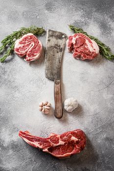 Various beef Delmonico steak or Ribeye Steak, Bone-In, with chuck eye roll cut with the meat american cleaver knife over grey stone table slate cutting top view normal face styled art with seasonings, tomatoes, garlic, rosemary.