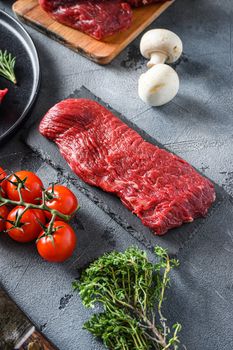 Raw tri tip, bottom sirloin cut steak on black slate raw organic food , marbled beef over other alternative cuts with herbs tomatoes peppercorns over grey stone surface background side view vertical.