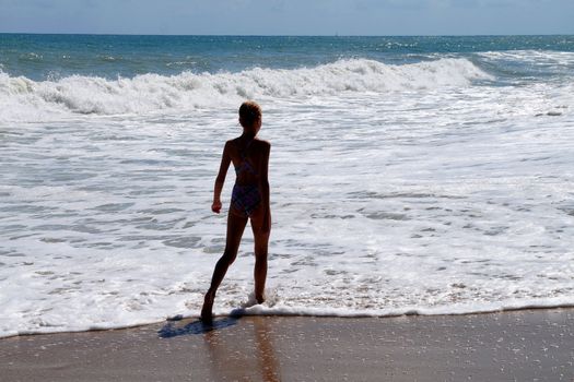 teenage girl walking alone along the empty seashore on the sunny day, rear view.