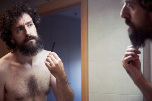 Hispanic middle-aged, curly hair and bare torso, fixes his long beard with a razor