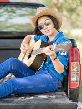 Young asian women short hair wear hat and sunglasses playing guitar ,sit on pickup truck in countryside Thailand