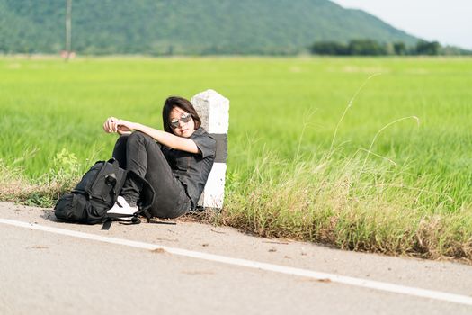 Young asian woman short hair and wearing sunglasses sit with backpack hitchhiking along a road wait for help in countryside Thailand