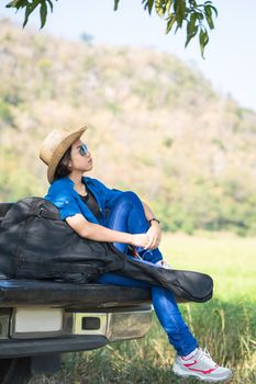 Young asian women short hair wear hat and sunglasses carry her guitar bag ,sit on pickup truck in countryside Thailand