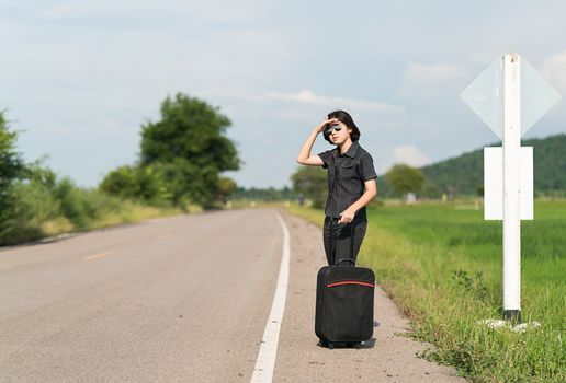 Young asian woman short hair and wearing sunglasses with luggage hitchhiking along a road in countryside Thailand