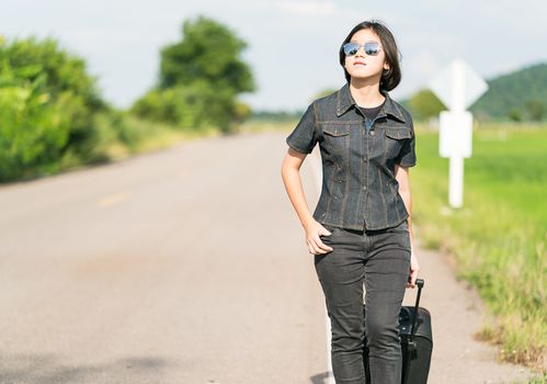 Young asian woman short hair and wearing sunglasses with luggage hitchhiking along a road in countryside Thailand