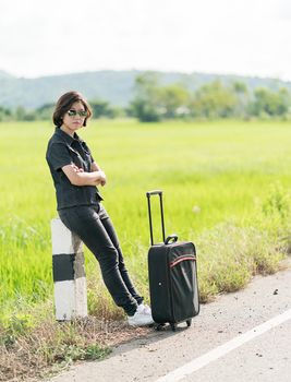 Young asian woman short hair and wearing sunglasses with luggage hitchhiking along a road in countryside Thailand