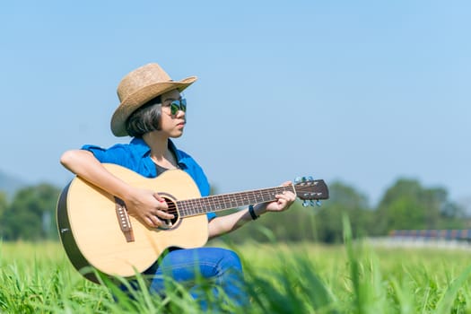 Young asian women short hair wear hat and sunglasses sit playing guitar in grass field countryside Thailand