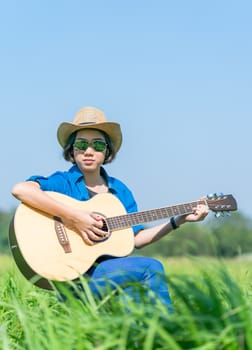 Young asian women short hair wear hat and sunglasses sit playing guitar in grass field countryside Thailand