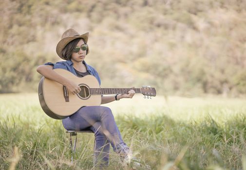 Young asian women short hair wear hat and sunglasses sit playing guitar in grass field countryside Thailand