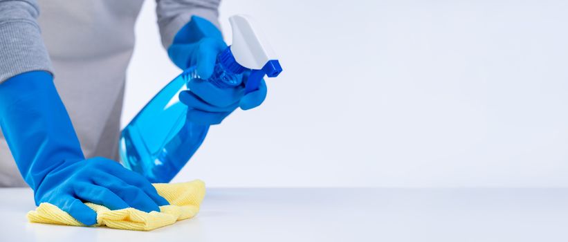 Young woman housekeeper in apron is cleaning, wiping down table surface with blue gloves, wet yellow rag, spraying bottle cleaner, closeup design concept.