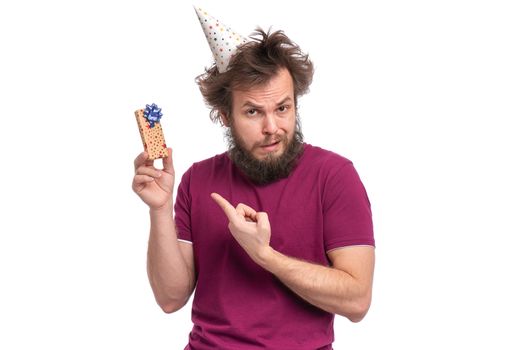 Crazy bearded Man with funny Haircut in birthday cap holding small gift box, isolated on white background. Surprised sad guy dissatisfied with his present. Holidays concept.