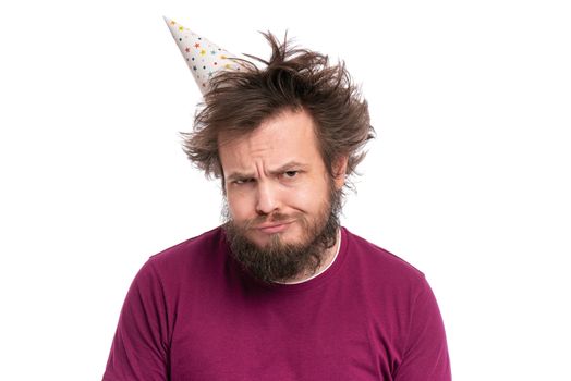 Crazy bearded Man with funny Haircut in birthday cap, isolated on white background. Unappy guy makes Offended Upset grimace. Holidays, Emotions and Signs concept.