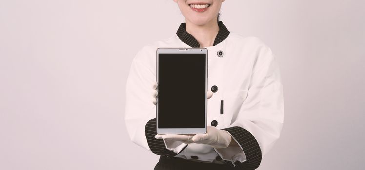 Middle aged of asian woman chef holding smartphone or digital tablet and received order from online shop or merchant application. she smiling in chef uniform and standing in studio with white color background.