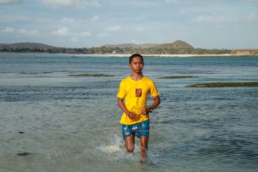 Topless, athletic and healthy black man running along the beach, splashing water during sunset. Teenager in a yellow T-shirt running in shallow water. Young man in shorts trains on the beach.