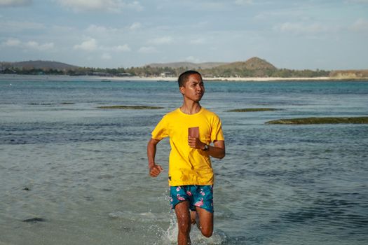 running asian man in water of tropical sea. Handsome man with athletic body running around the beach line near the sea.Fit runner on the beach with sea waves background. Beach activities concept.