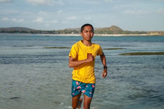 running asian man in water of tropical sea. Handsome man with athletic body running around the beach line near the sea.Fit runner on the beach with sea waves background. Beach activities concept.