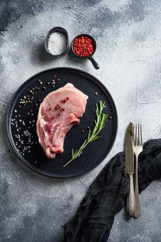 pork meat, cutlet in a black round plate on a grey textured stone background with fork and knife rosemary garlic peppercorns ingredients top view vertical.