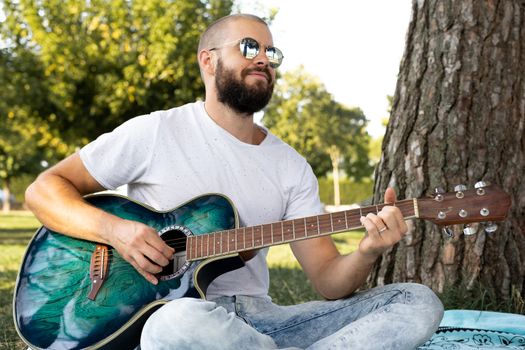 portrait of a young shaved and bearded man playing guitar in the park