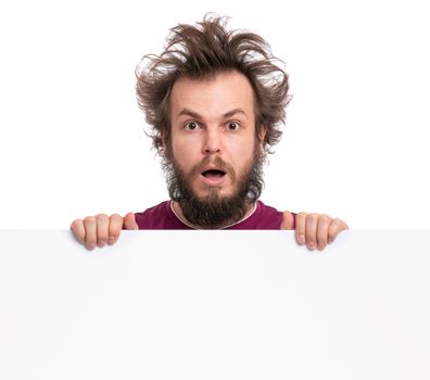 Crazy bearded Man with funny Haircut showing empty blank signboard with copy space. Guy with surprised eyes and mouth open peeking out from behind big white banner, isolated on white background.