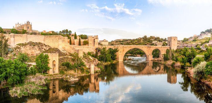 Beautiful landscape of Toledo in Spain. Stone bridge across calm river. Blue sky reflected in crystal clear water. Big fort and country houses in background. Popular tourist place in Europe.