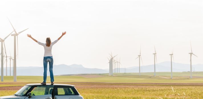 Young woman standing on top of her car with hands raised. Girl in yellow fall field with wind turbines in background. Alternative energy source. Power generation. Ecology and freedom concept.