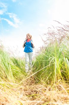 Happy smiling woman walking in field. Green grass in foreground and clear sky in background. Girl in sport clothes hiking with backpack. Blonde crossing meadow. Outdoor activity and active lifestyle.