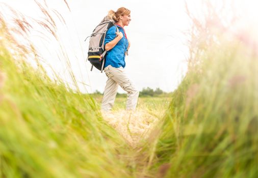 Happy smiling woman walking in field. Green grass in foreground and clear sky in background. Girl in sport clothes hiking with backpack. Blonde crossing meadow. Outdoor activity and active lifestyle.