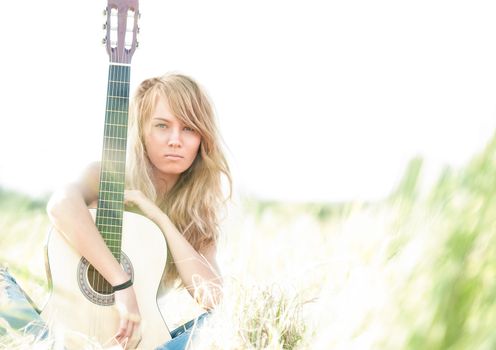 Pretty girl with guitar sitting on ground. Beautiful woman with blonde hair holding musical instrument and smiling. Green grass as background. Sunny summer day. Outdoor activity for young people.