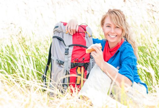 Young pretty woman tourist with backpack having lunch in wheat field background. Happy and smiling girl on halt. Tourism travelling and hiking outdoors in summer. Healthy lifestyle.