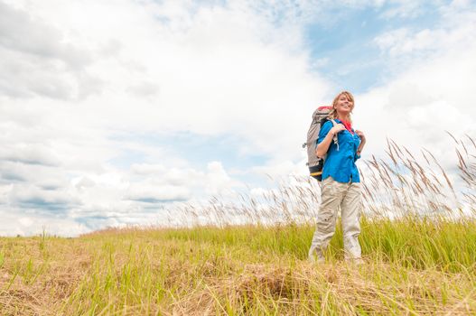 Happy smiling woman walking in field. Green grass in foreground and clear sky in background. Girl in sport clothes hiking with backpack. Blonde crossing meadow. Outdoor activity and active lifestyle.